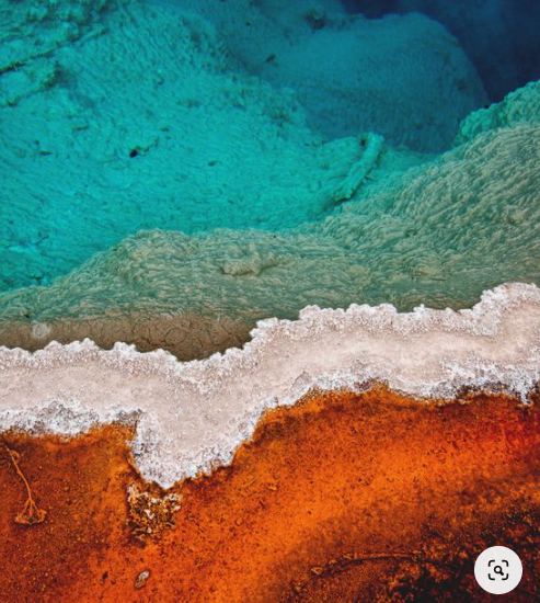 Arial image of a sea coast, with turquoise water, soft peach sand, and bright orange landforms.