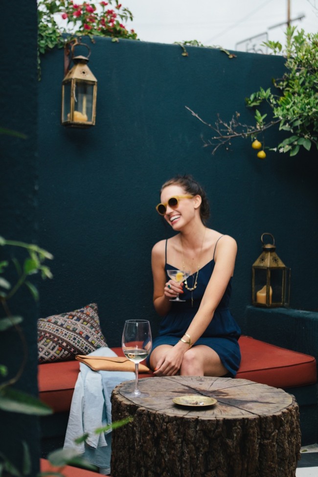 A lifestyle image of a woman in a navy dress, sitting against a dark teal wall, on a red cushion, in a garden with a lemon tree.